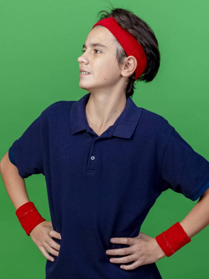 confident young handsome sporty boy wearing headband and wristbands with dental braces keeping hands on waist looking at side isolated on green background with copy space